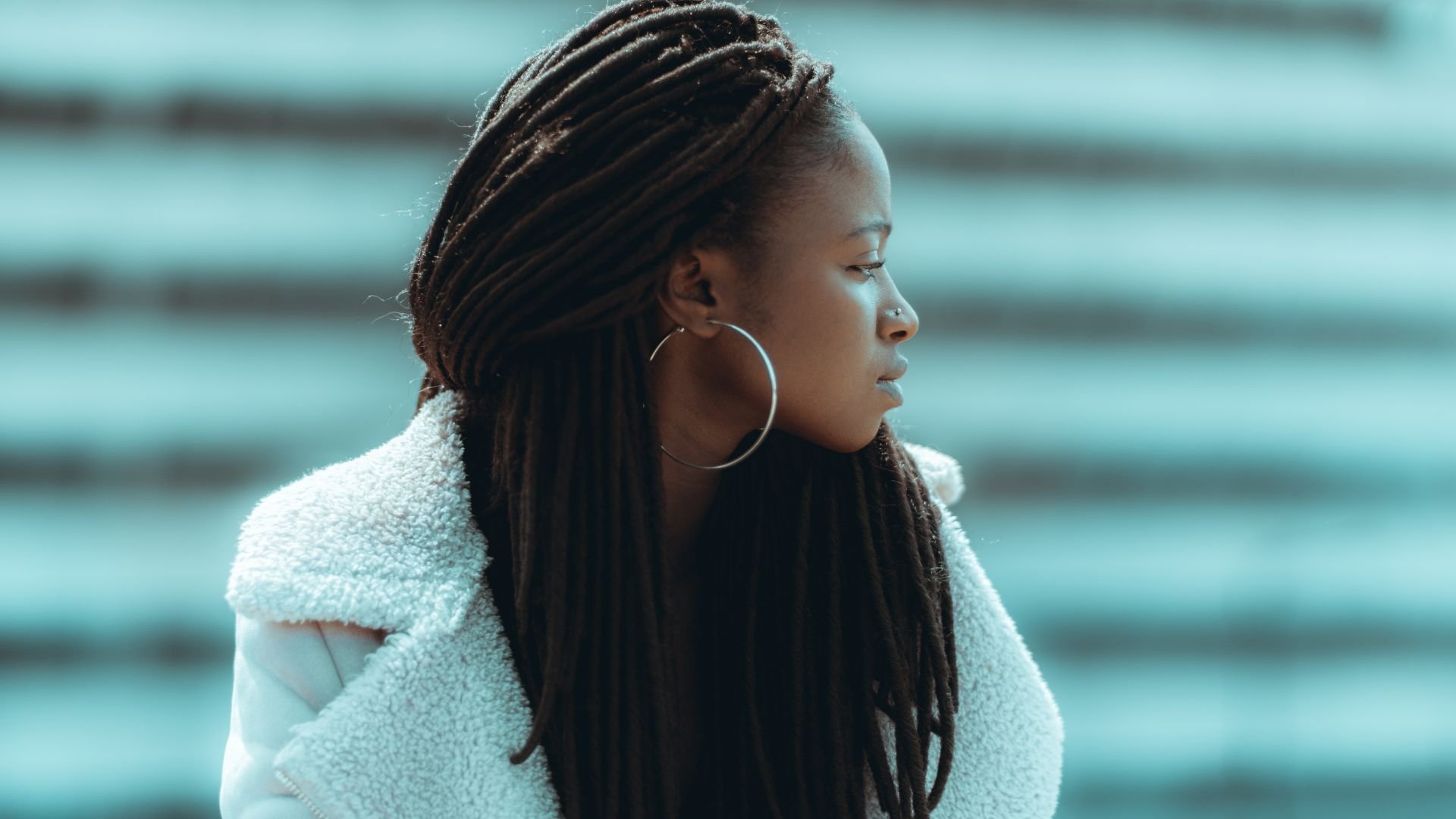 Person with long braids in white coat looking sideways against blue background